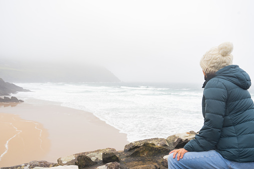 Unknown woman with a knit hat an jacket spotting an exceptional view of a wild beach on the Irish coast on a foggy and windy day