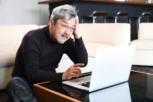 An older Asian man with gray hair is having trouble operating a laptop computer.