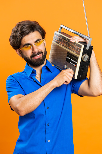 Man in sunglasses using retro tape record player to listen music, disco dancing of favorite track, having fun, entertaining, fan of vintage technologies. Arabian guy isolated on orange background