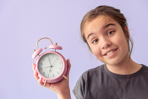 Teenage girl with desk alarm clock. Hands on the clock show 7, Close-up portrait, purple background