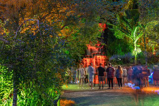 A group of people stand in front of the illuminated waterfall as the colours change. The majority of the people are blurred due to the long exposure. Light streaks from children wielding light sabers add to the colours, as do the lights illuminating the surrounding vegetations.