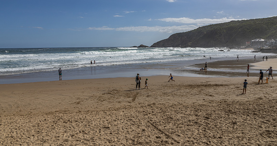 People enjoy a warm spring day at Herolds Bay near George in the Western Cape. A group of people is playing cricket, while others swim or paddle. One boy is chasing the cricket ball, while a man with a bat stands on the sand by the stumps.