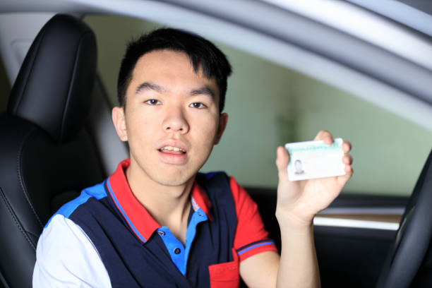 A teenage male Asian new learner driver is holding up his drivers license. He is smiling at the camera. He is wearing a colorblocked golf shirt.