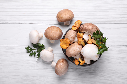 Bowl with different mushrooms and parsley on white wooden table, flat lay