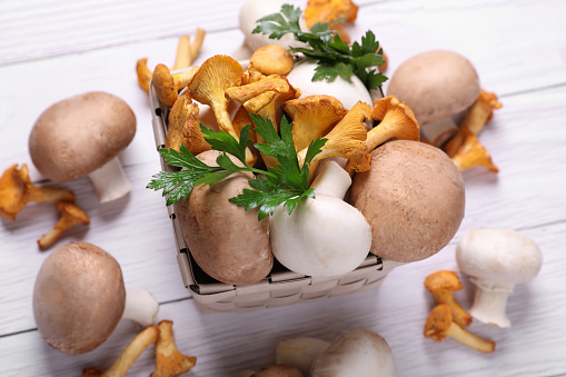 Basket with different mushrooms and parsley on white wooden table, above view
