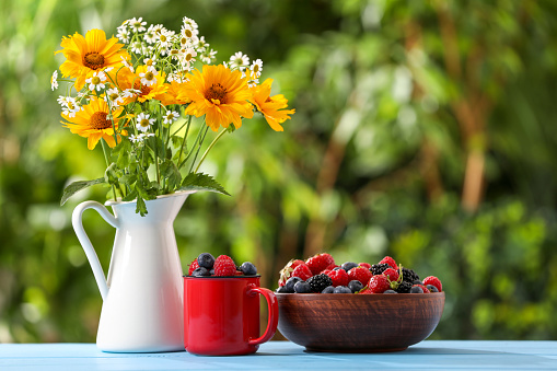 Different fresh ripe berries and beautiful flowers on light blue table outdoors