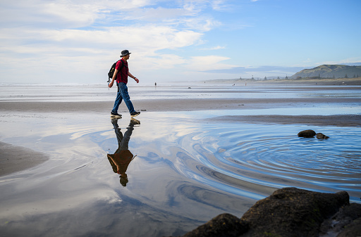 Man walking with reflection on wet sand at Muriwai Beach in summer. Auckland.