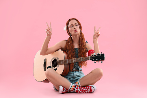 Stylish young hippie woman with guitar showing V-sign and sending air kiss on pink background