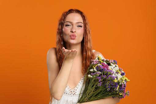 Beautiful young hippie woman with bouquet of colorful flowers blowing kiss on orange background