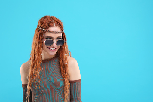 Stylish young hippie woman in sunglasses on light blue background, space for text