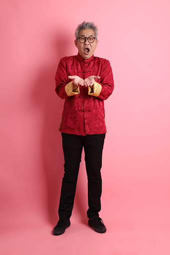 Happy Chinese new year. Asian Chinese energetic senior man wearing red traditional cheongsam qipao or changshan dress with gesture of hands giving isolated on pink background.