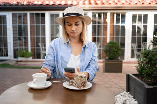Young woman wearing summer hat typing text message on smart phone in a cafe. Girl with a coffee and dessert using mobile phone in coffee shop. Copy space
