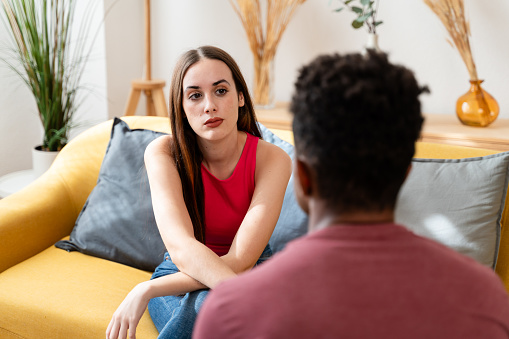 Sad young woman sitting on sofa and talking to her boyfriend at home
