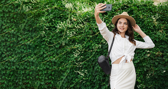 Smiling girl in straw hat takes selfie, video chats, holds smartphone with camera, posing in summer park on green background. Copy space