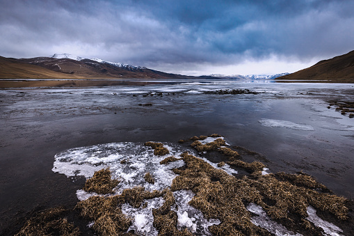 A serene winter landscape captured at Lake Tso Moriri in Ladakh, India. The frozen lake reflects a clear blue sky adorned with heavy, fluffy clouds while showcasing ice formations along the shore with distant mountain vistas.