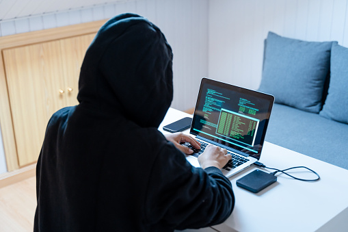 Hacker in black hoodie stealing information on laptop at home.