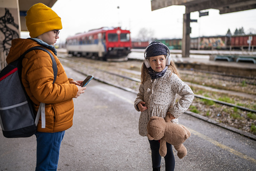 Cute brother and sister standing on train station, girl holding Teddy bear while boy using mobile phone
