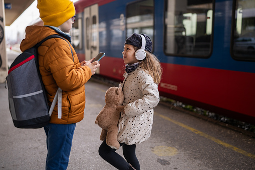 Cute brother and sister standing on train station, girl holding a toy while boy using mobile phone