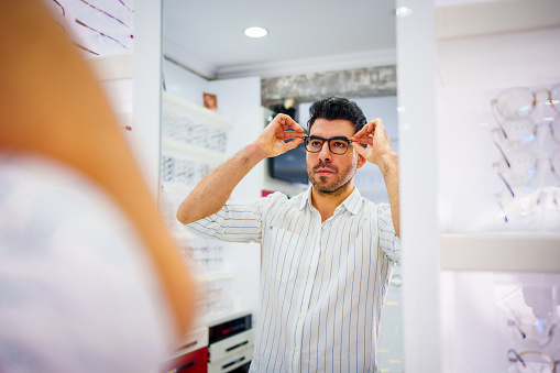 Smiling young man trying on glasses on mirror in optician