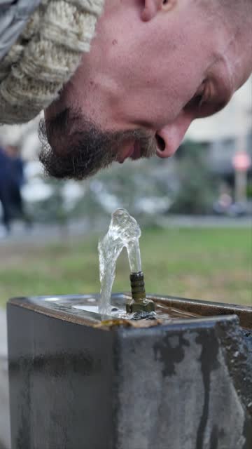 Vertical video of a young bearded man bending over drinking clean drinking water in a special fountain in a public park. A man uses pulpulak to quench his thirst by drinking water.