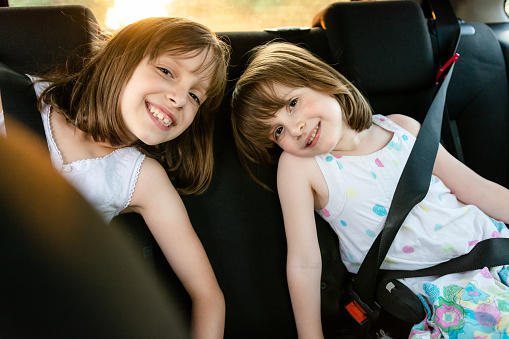Two happy girls are sitting on the back seat of the car and looking at the camera.