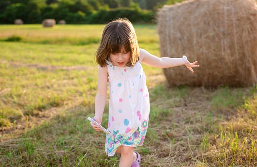 A little girl in a summer dress walks through a meadow with hay bales on a summer day.