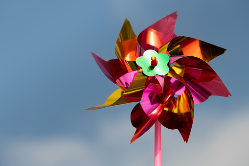 A colourful windmill for children spins in front of a blue sky with clouds. The sun is shining. The toy shines.