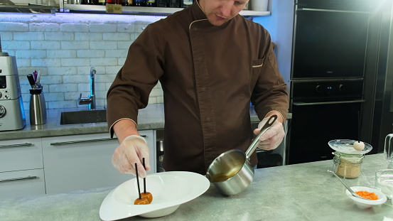 Chef in jacket places candy in milted caramel on plate. Man performs high-level presentation of dessert in kitchen restaurant