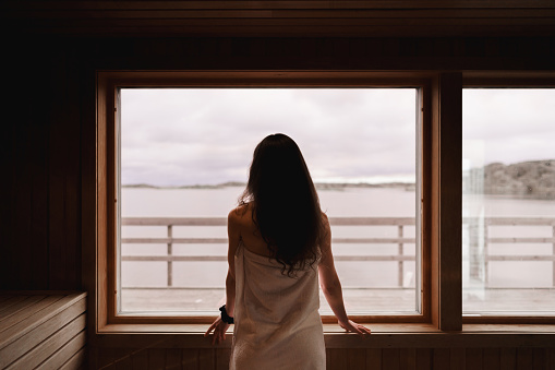 Photograph of the inside of an empty dry sauna with large windows. Walls and benches made of wood and a tile floor. A woman is standing by the large window, looking out at the view of the Swedish west coast.
