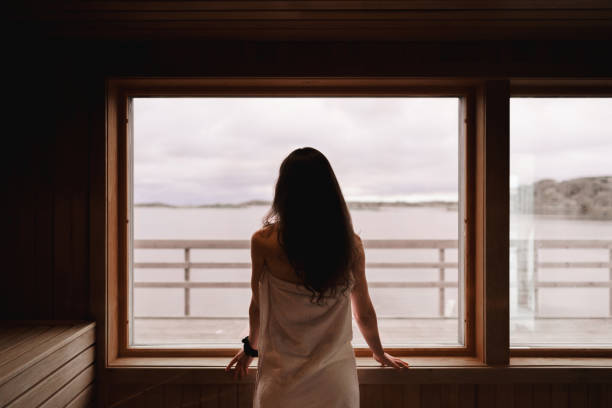 Woman wearing a towel is standing by the window in a dry sauna Photograph of the inside of an empty dry sauna with large windows. Walls and benches made of wood and a tile floor. A woman is standing by the large window, looking out at the view of the Swedish west coast. swedish sauna stock pictures, royalty-free photos & images