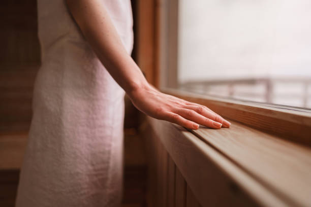 Photograph of the inside of an empty dry sauna with large windows. Walls and benches made of wood and a tile floor. A woman is standing by the large window, looking out at the view of the Swedish west coast.