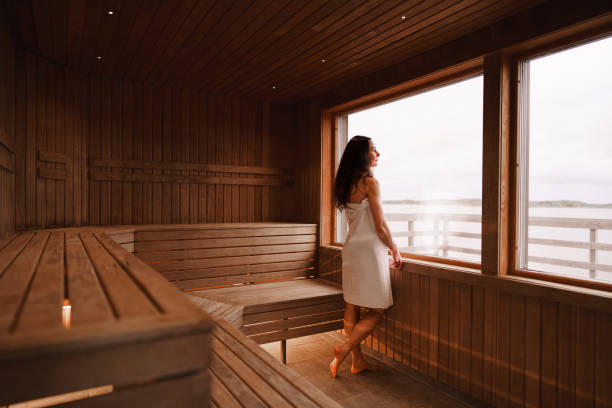 Woman wearing a towel is standing by the window in a dry sauna Photograph of the inside of an empty dry sauna with large windows. Walls and benches made of wood and a tile floor. A woman is standing by the large window, looking out at the view of the Swedish west coast. swedish sauna stock pictures, royalty-free photos & images