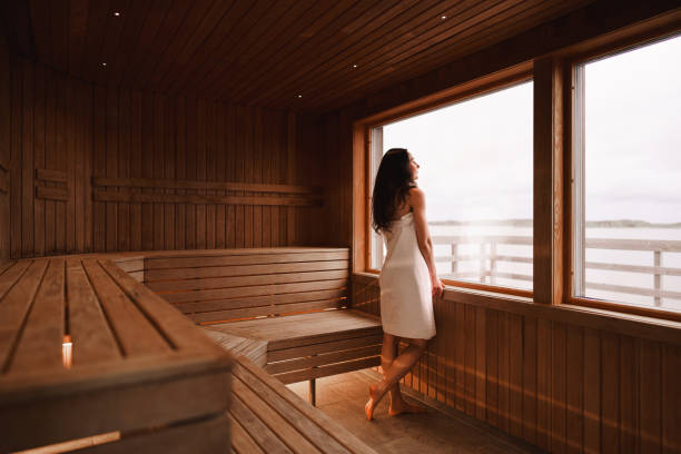 Woman enjoying the view from a dry sauna Photograph of the inside of an empty dry sauna with large windows. Walls and benches made of wood and a tile floor. A woman is standing by the large window, looking out at the view of the Swedish west coast. swedish sauna stock pictures, royalty-free photos & images