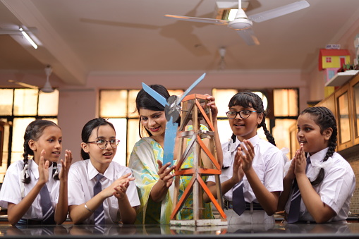 Secondary Or High School Students Studying Wind Turbines In Science Class With Teacher. Girls are excited to explore lab project. Electricity generate