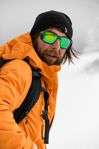 portrait of bearded man in bright jacket and black hat with glasses with reflection of winter ski resort.