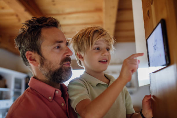 Boy helping father to adjust, lower heating temperature on thermostat. Concept of sustainable, efficient, and smart technology in home heating and thermostats. Young boy helping father to adjust, lower heating temperature on thermostat. Concept of sustainable, efficient, and smart technology in home heating and thermostats. smart thermostat stock pictures, royalty-free photos & images