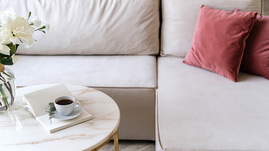 Selective focus on marble table with black coffee in with cup. Open book or diary journal with copy space paper pages close to vase with flowers next to comfortable couch with cushions in living room