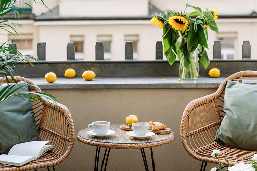 Round coffee table close to two rattan armchairs with cushions at open balcony. Hot tea drink in cups and croissant. Good morning concept. Breakfast in cozy hotel patio with flowers.