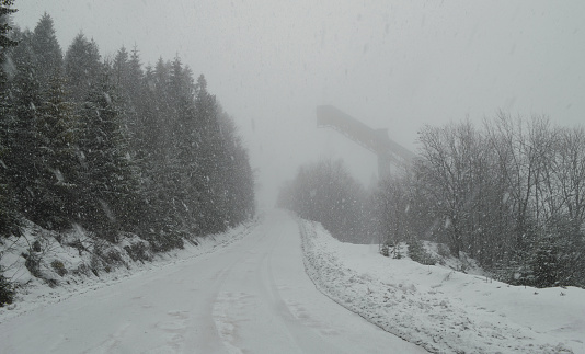 Heavy snow falls on a coniferous forest and a road going off into the distance. Abandoned old ski jump in the background. Monochrome winter landscape.