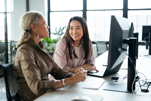 Businesswomen in 30s and 50s sitting in front of desktop computer and interacting while working on project.