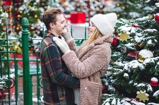 Portrait of young loving happy beautiful couple having fun enjoying decorated Xmas market. Kiss, hug, embrace each other, precious magic moments together on holiday time. Warm clothes, snow
