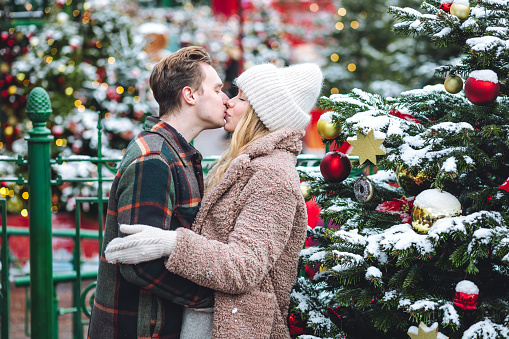 Beautiful young loving couple, boyfriend and girlfriend having fun on a Christmas market wearing warm clothes, taking pictures for family, social network. Outdoors, winter time, snowy weather.