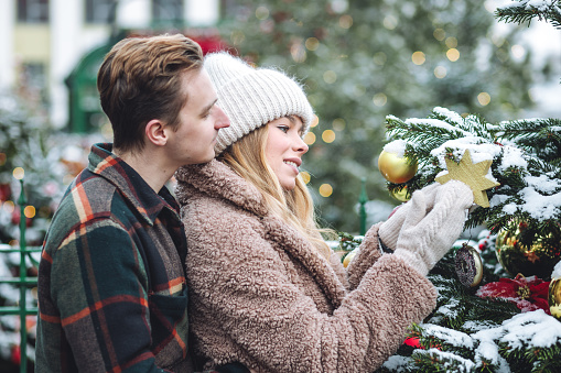Portrait of young loving happy beautiful couple having fun enjoying decorated Xmas market. Kiss, hug, embrace each other, precious magic moments together on holiday time. Warm clothes, snow