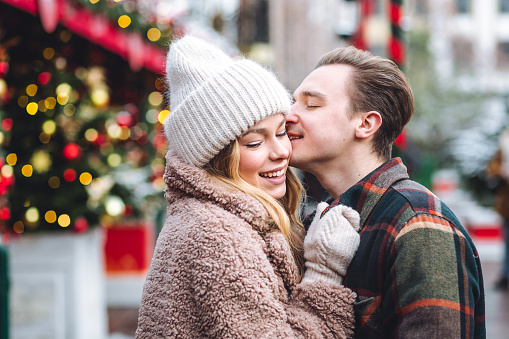 Beautiful young loving couple, boyfriend and girlfriend having fun on a Christmas market wearing warm clothes. Outdoors, winter time, snowy weather. Tangerines in string bag, fir tree branches