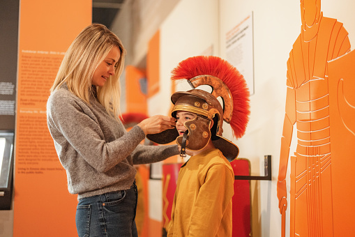 An attractive caucasian mother letting her son try a Roman galea during their visit of the history museum. The mother is putting on the Roman helmet while the son is standing still.  The young boy looks very happy and is smiling while his mom looks focused.