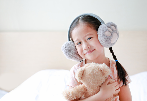 Smiling little Asian child girl wearing winter earmuffs and embracing teddy bear while sitting on the bed at home.