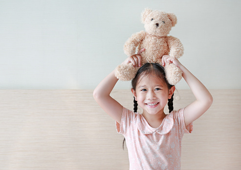 Smiling Asian little girl showing teddy bears while sitting on the bed at home.