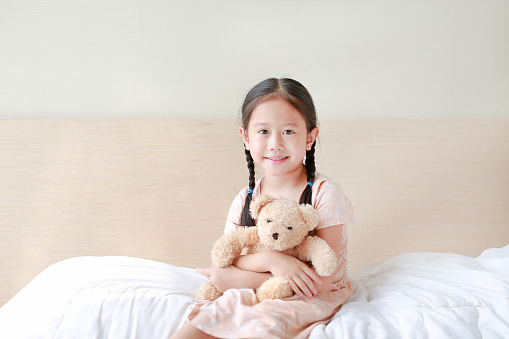 Smiling Asian little girl cuddle teddy bear and looking camera while sitting on the bed at home.