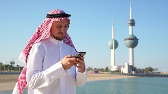 Arab Man In Traditional Arabic Dishdasha Using His Phone In Front Of Kuwait Towers