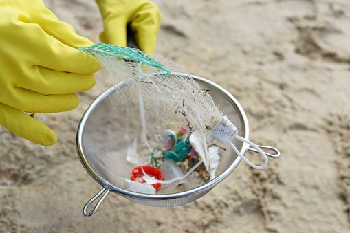 The close-up image of a volunteer wearing glove and using colander to clear away plastic pieces and rubbish on the beach. Volunteers play a crucial role in helping to reduce the impact of plastic pollution on coastal ecosystems.